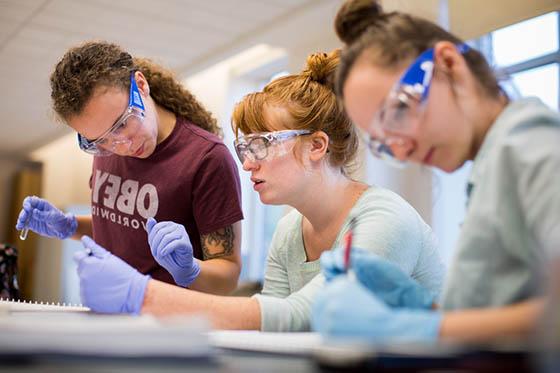 图为三名足球外围买球靠谱的女学生戴着护目镜和手套在实验室工作